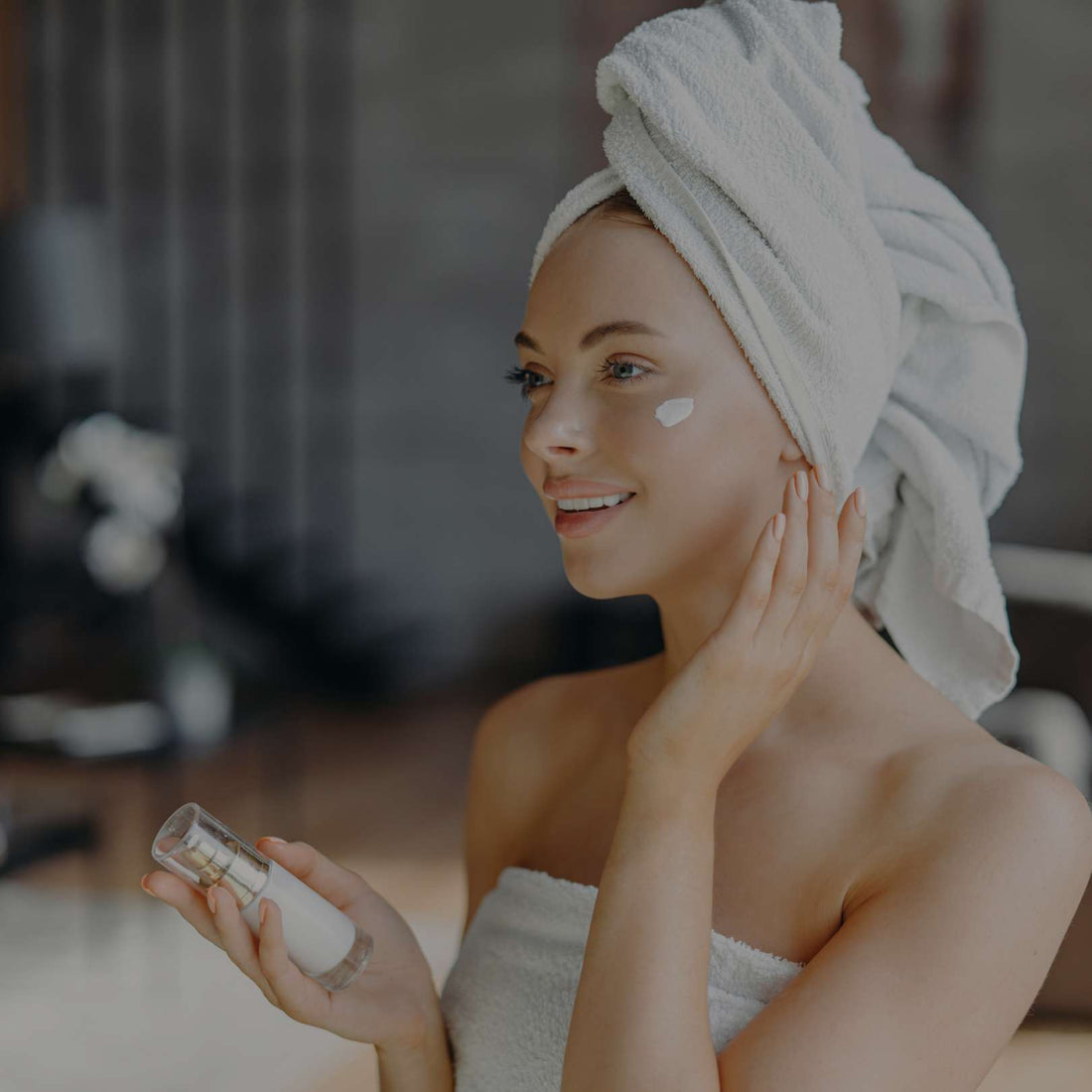 Woman with a towel wrapped around her head applying facial moisturizer while holding a skincare bottle during her daily beauty routine.