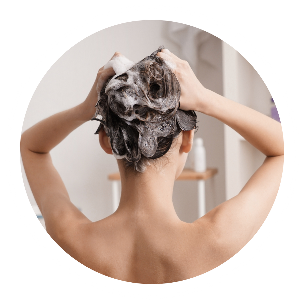 A person viewed from behind with their head covered in lathered shampoo, arms raised as they wash their hair, centered inside a circular frame on a white background.