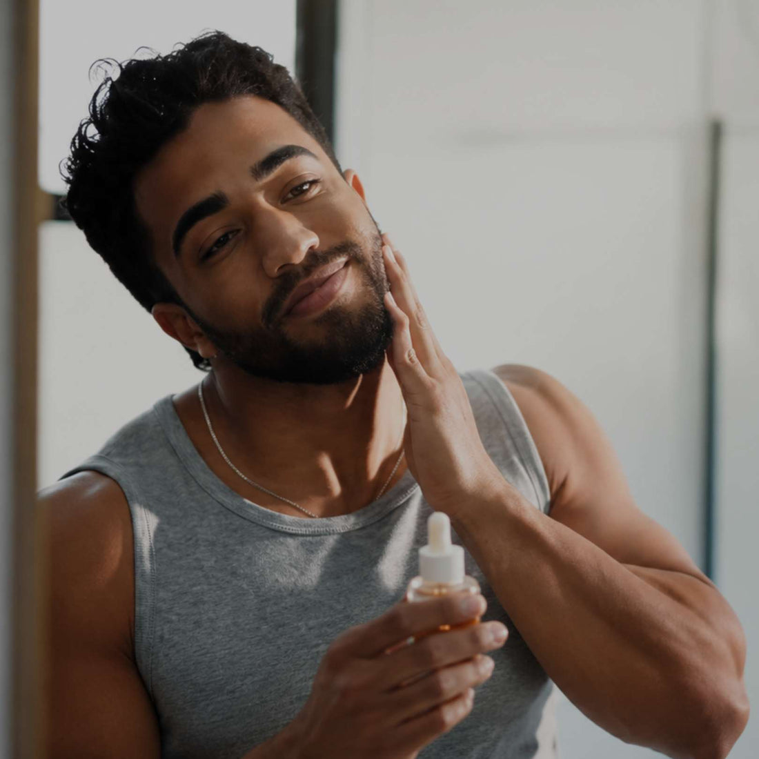 A man in a gray tank top applies a skincare serum from a small dropper bottle to his face, captured in a bright indoor setting.