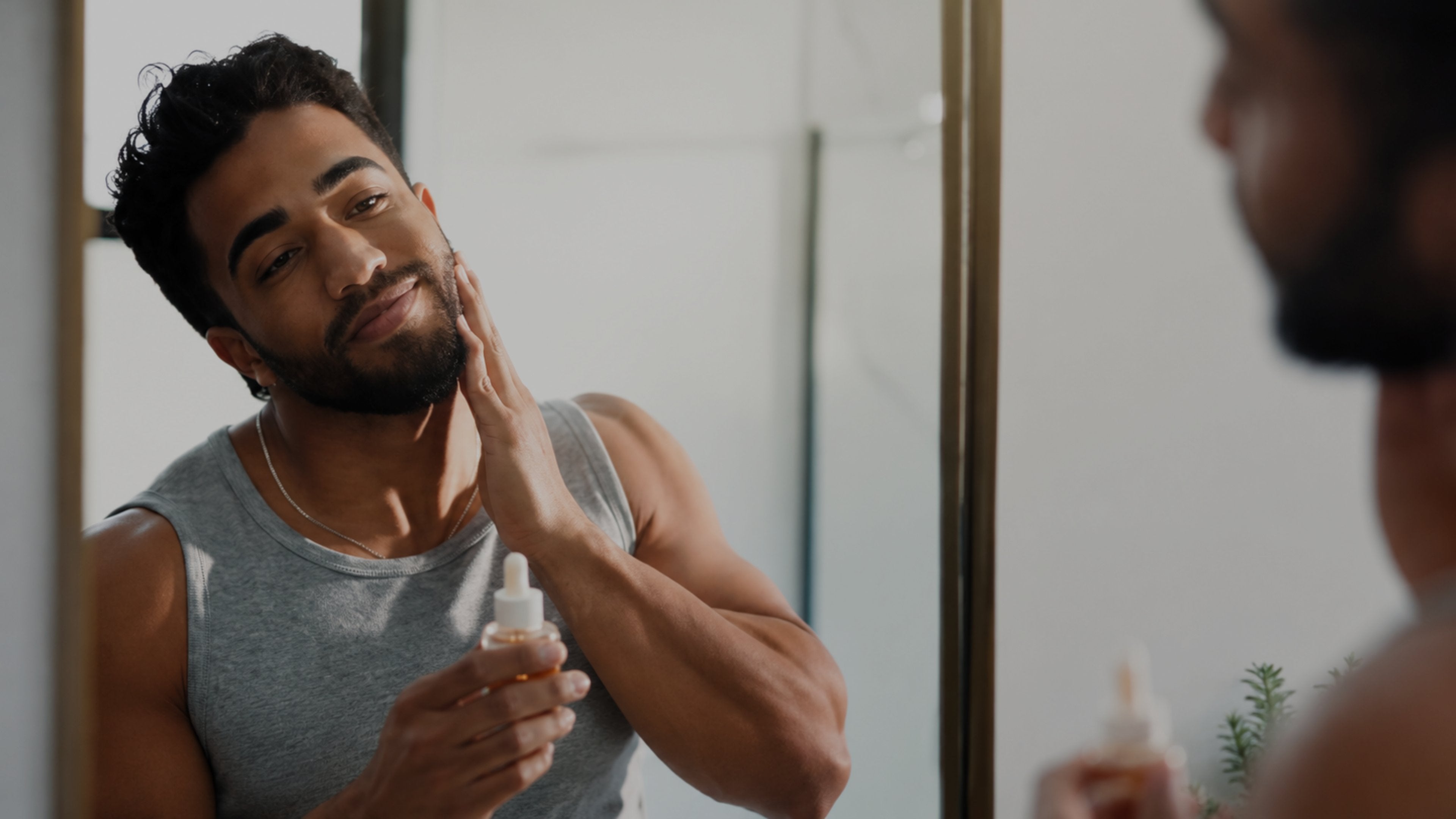 A man applies skincare cream from a small white bottle to his face while looking into a mirror in a bathroom.
