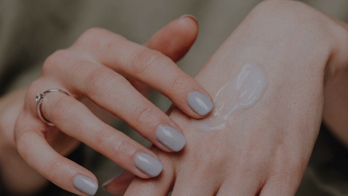 A close-up of a hand with light gray nail polish touching a clear, glossy cosmetic cream on the back of another hand.