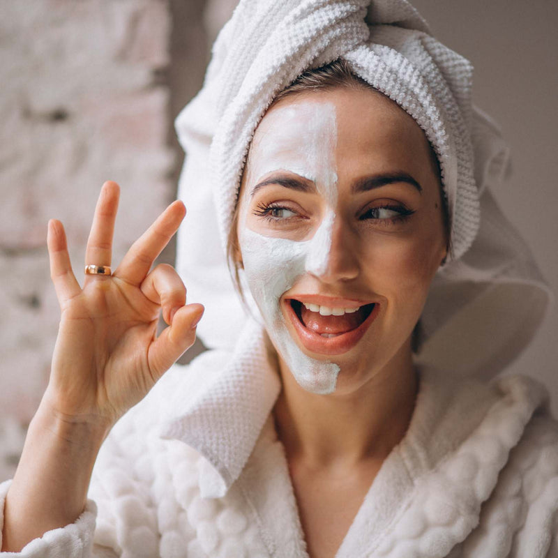 A smiling woman with a white face mask on one side of her face, wearing a white textured bathrobe and a white towel-wrapped hair, making an 'OK' hand gesture against a soft neutral background.