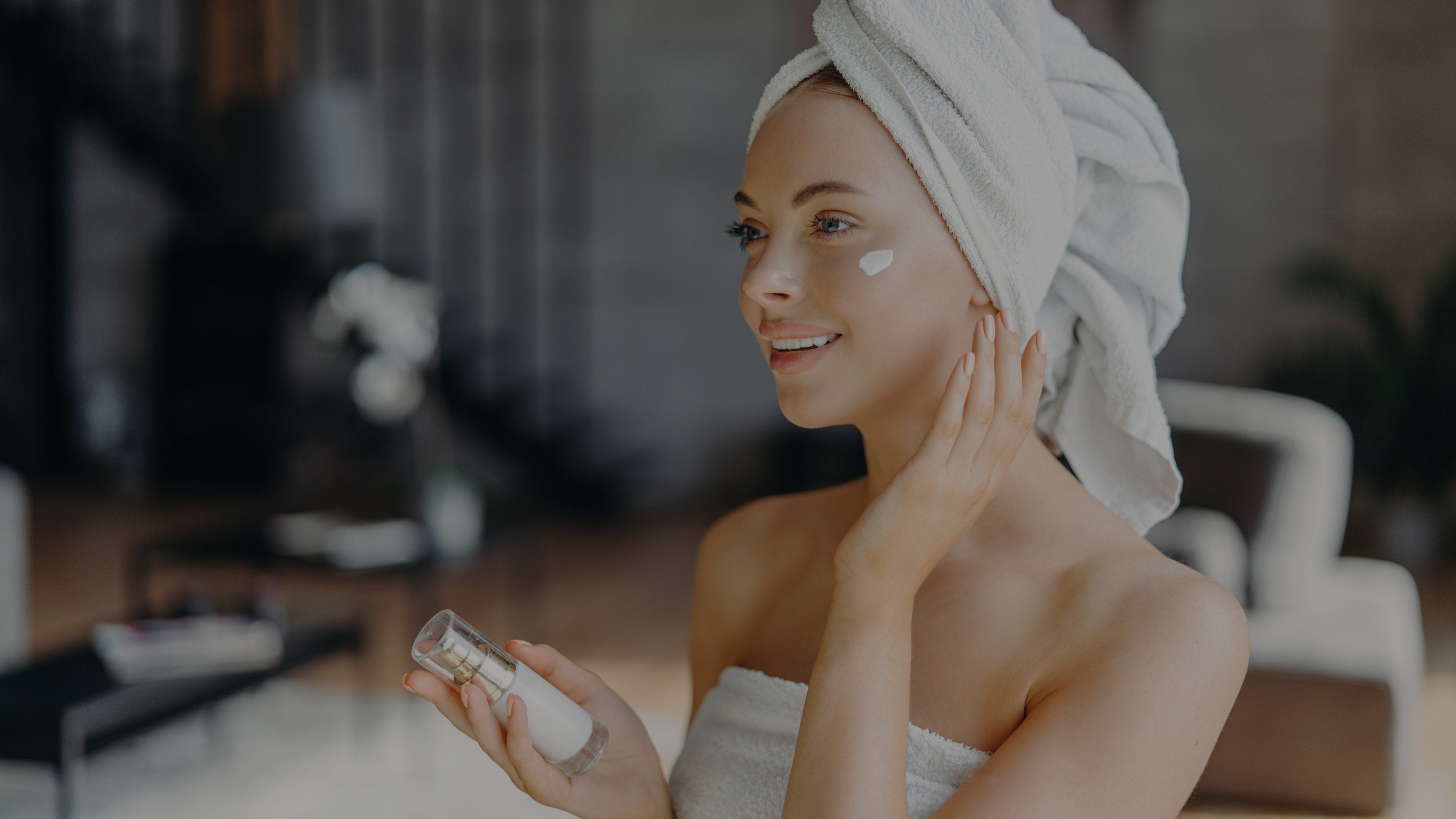 A woman with a white towel wrapped around her head applies cream to her cheek while holding a small cosmetic bottle in a softly lit indoor setting.